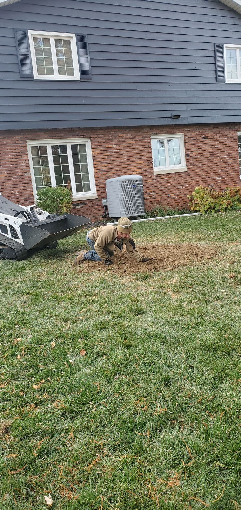 Child digging in the yard near a small excavator and a house with brick and siding.