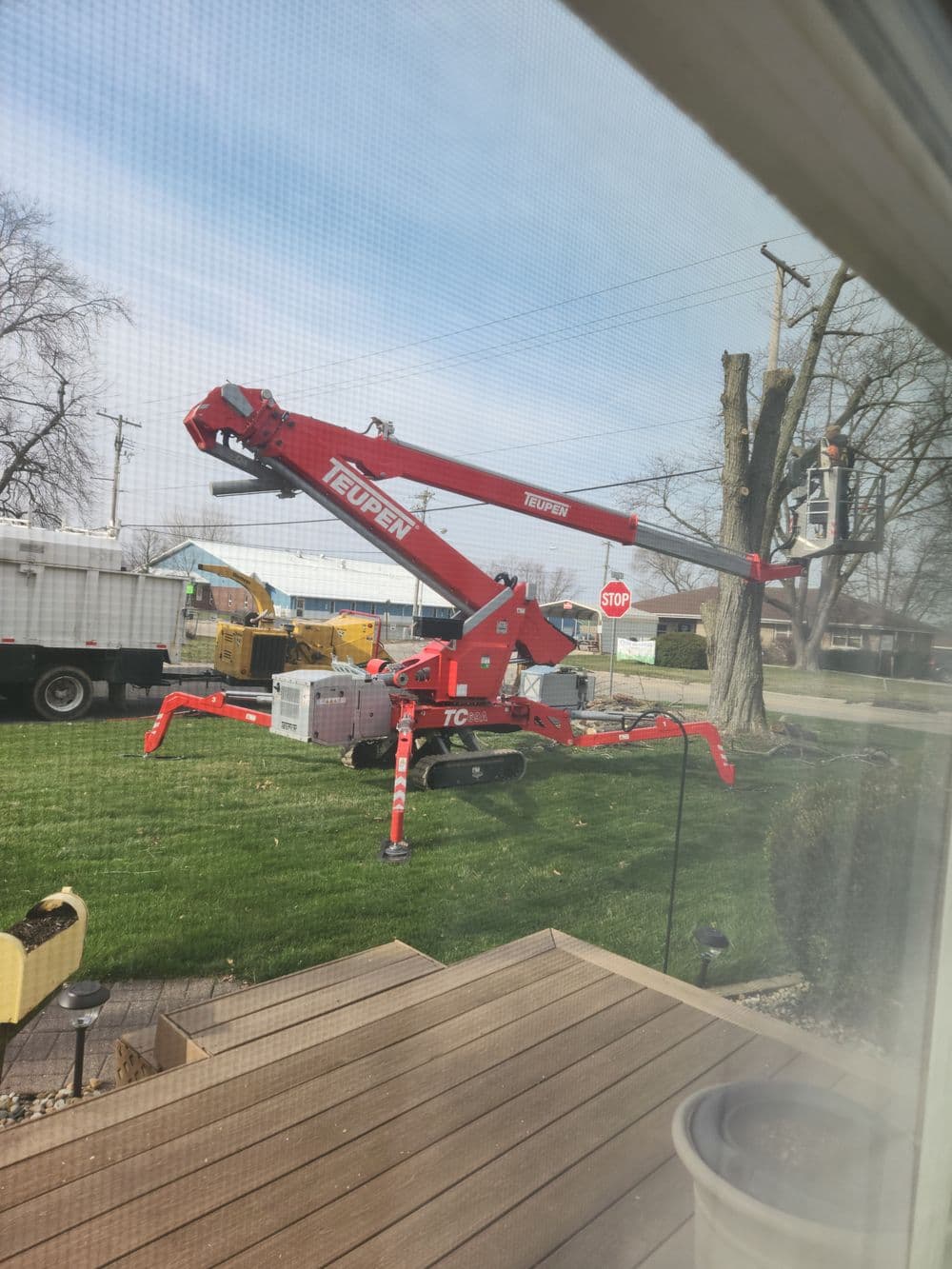 Red Teupen lift parked on lawn, tree work underway, with construction equipment in background.