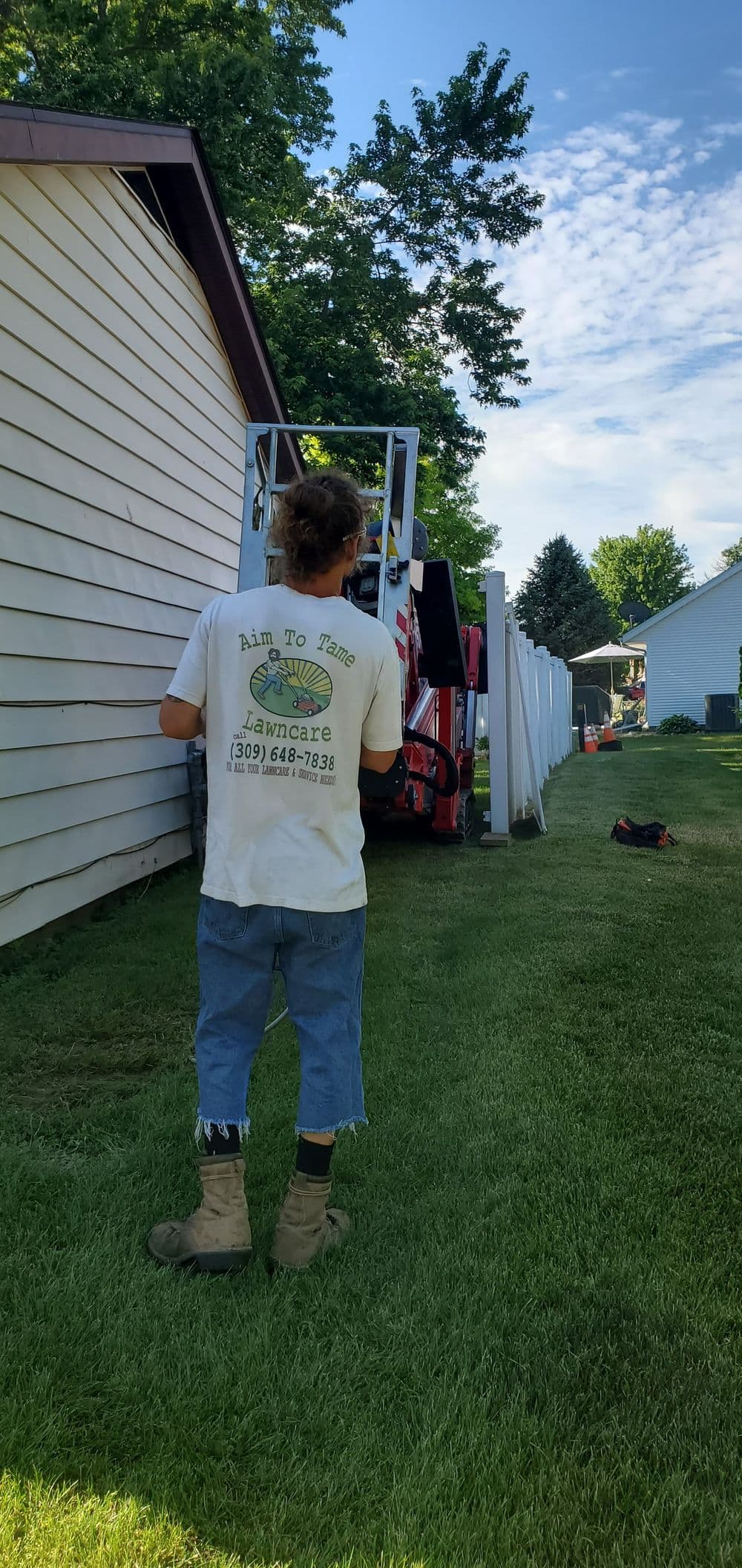 Worker operating lawn care equipment near a house, surrounded by green grass and trees.