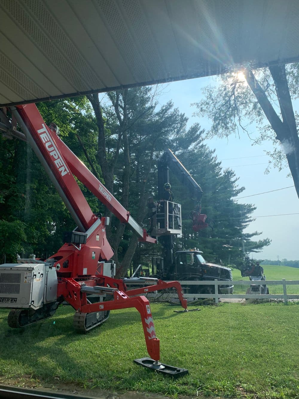 Red crane lifting workers near trees, with a clear sky and grassy area in view.