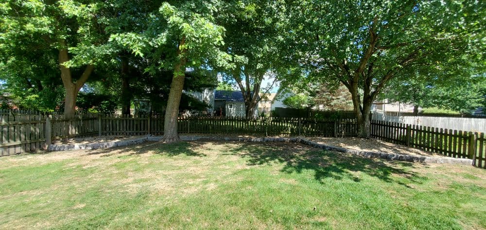 Lush backyard with trees, a stone border, and a wooden fence surrounding a grassy area.